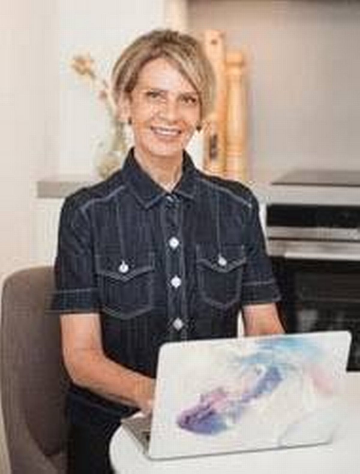 An adult woman with short blonde hair, wearing a navy blue shirt, stands smiling in a modern kitchen setting. The background includes a white wall adorned with a decorative item and a kitchen appliance partially visible.