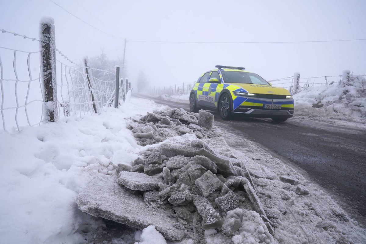 A vehicle navigates through a snowy road, with scattered debris and mounds of snow lining the roadside. The scene is set against a backdrop of fog, indicating adverse weather conditions.