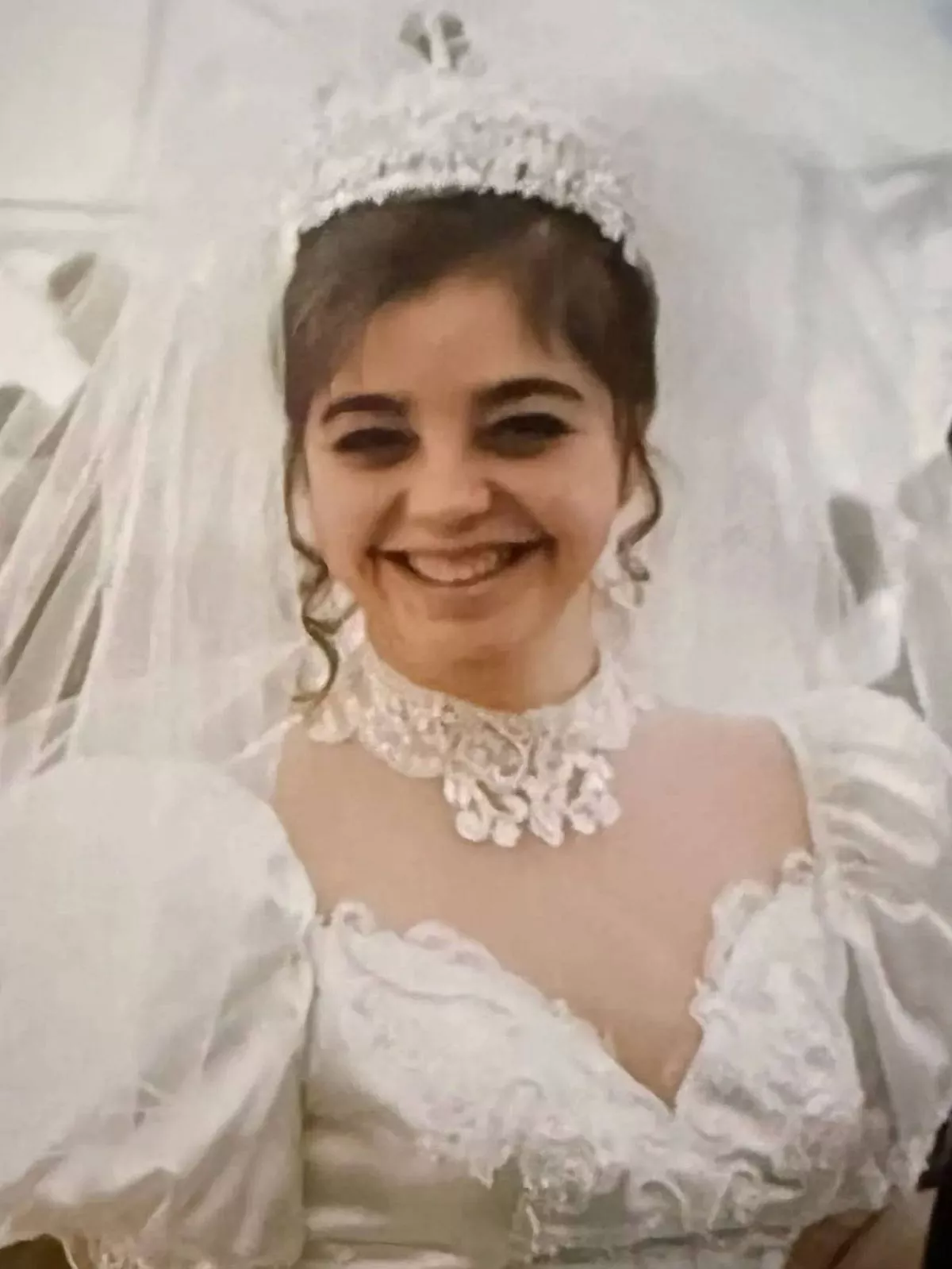 An individual dressed in a wedding gown, complete with a veil and a necklace, is captured in a portrait, exhibiting a joyful expression.