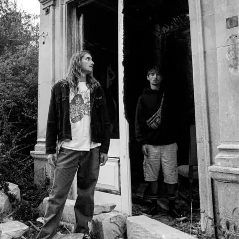 Torus Two young men photographed in a disused house, one stood on the left with long hair and one on the right with shorter hair and a light beard, shot in black and white