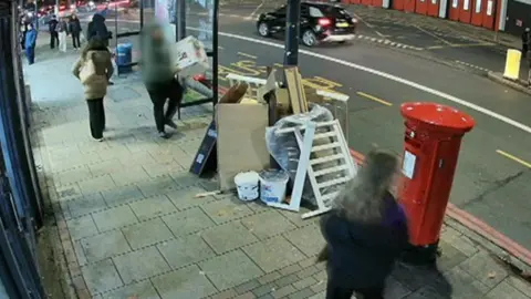 Handout CCTV still of a blurred person putting a large cardboard box on to a large pile of household wate next to a bus stop