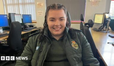 A young woman with dark hair in a pony tail is sat in a chair in the emergency call centre. She is wearing a dark green ambulance jacket and t-shirt, and is smiling to the camera.