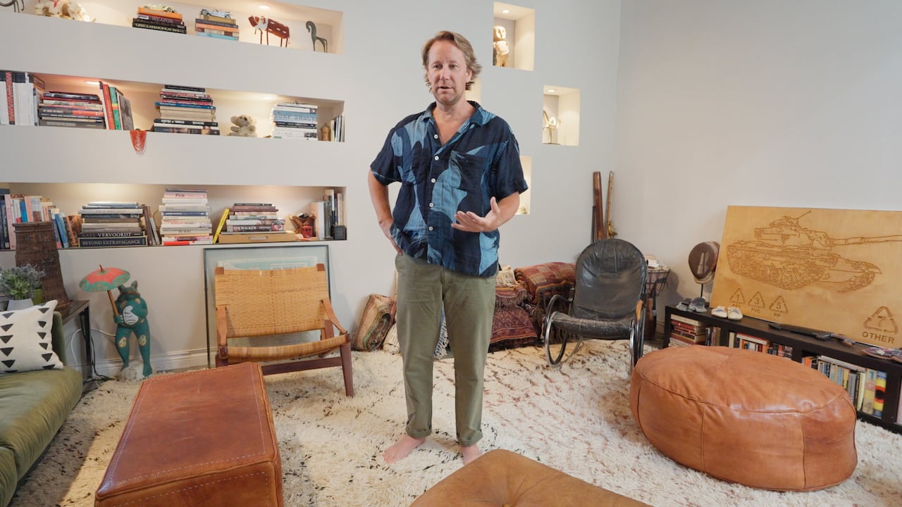 A man stands in his living room with a rug, low-to-the ground furniture and bookshelves. 