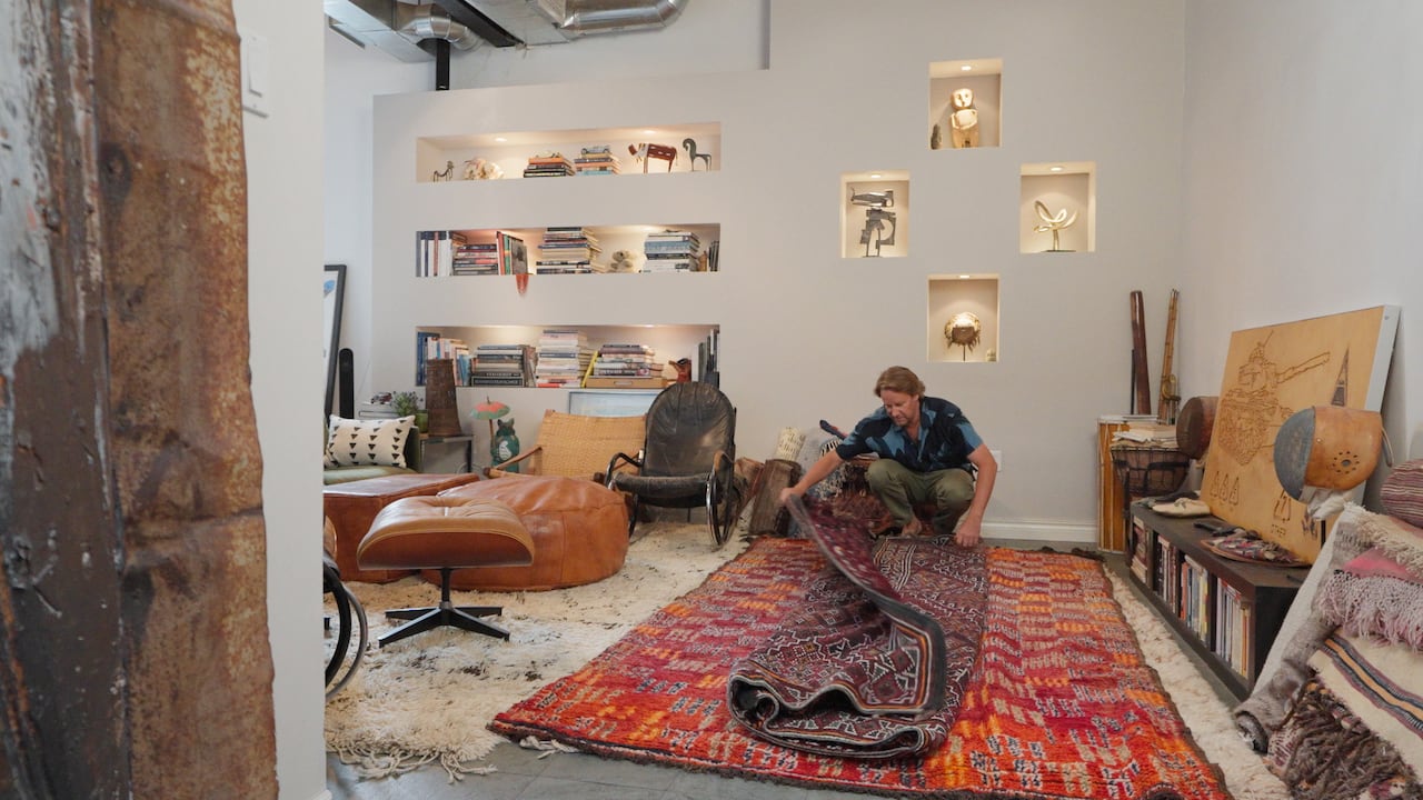 A man crouches, unrolling two rugs in his living room. 