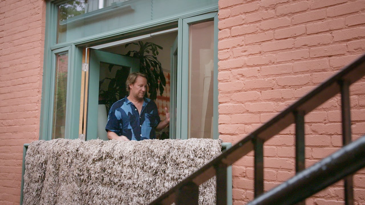 A man standing next to a Juliet balcony with a rug hanging over the railing. 