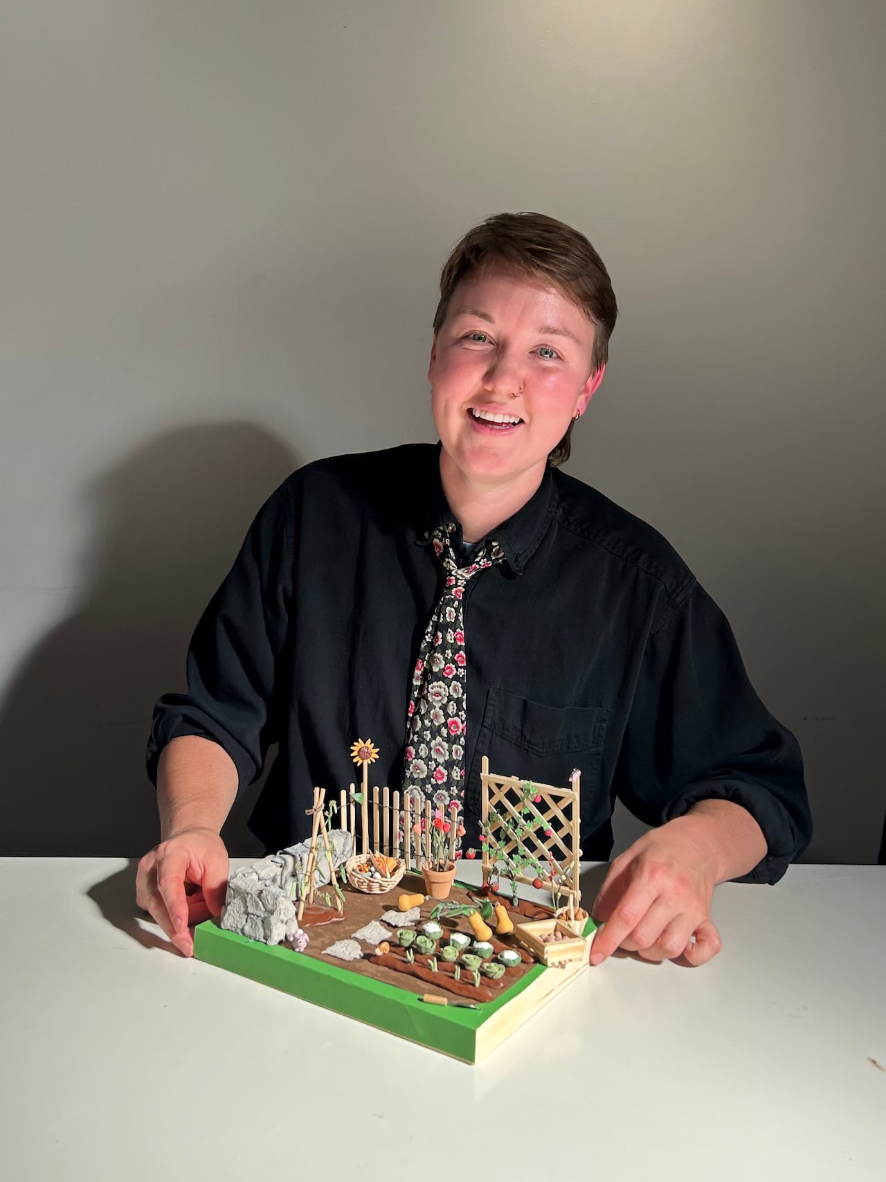 A person with short hair, a black shirt and a brightly coloured tie sits behind a miniature sculpture of a vegetable garden