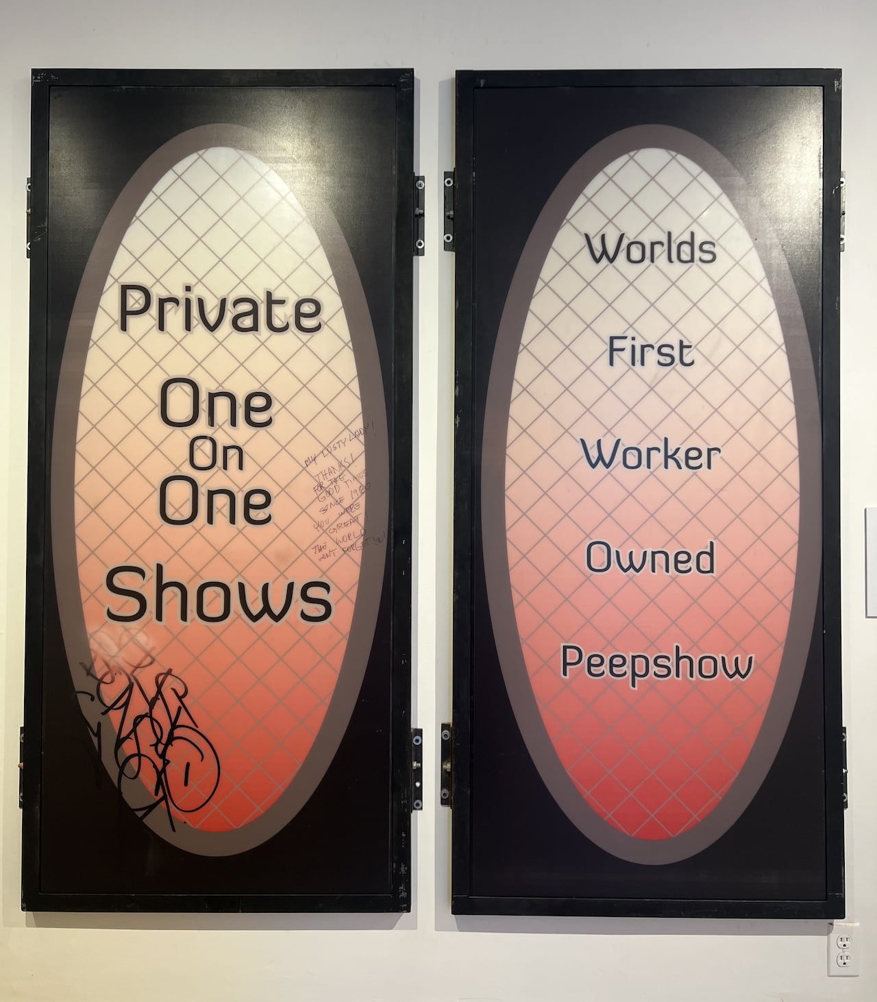 Hanging on a white wall, the lightbox signs show an oval opening with a chainlink motif. They read "Private one on one shows" and "Worlds first worker owned peepshow."