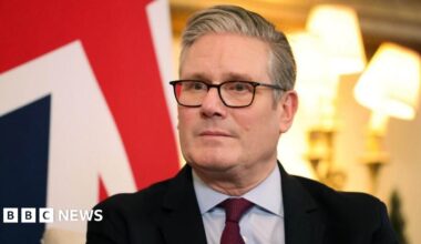 Headshot of Sir Keir Starmer in a suit sitting in front of two bright lamps and a UK flag.