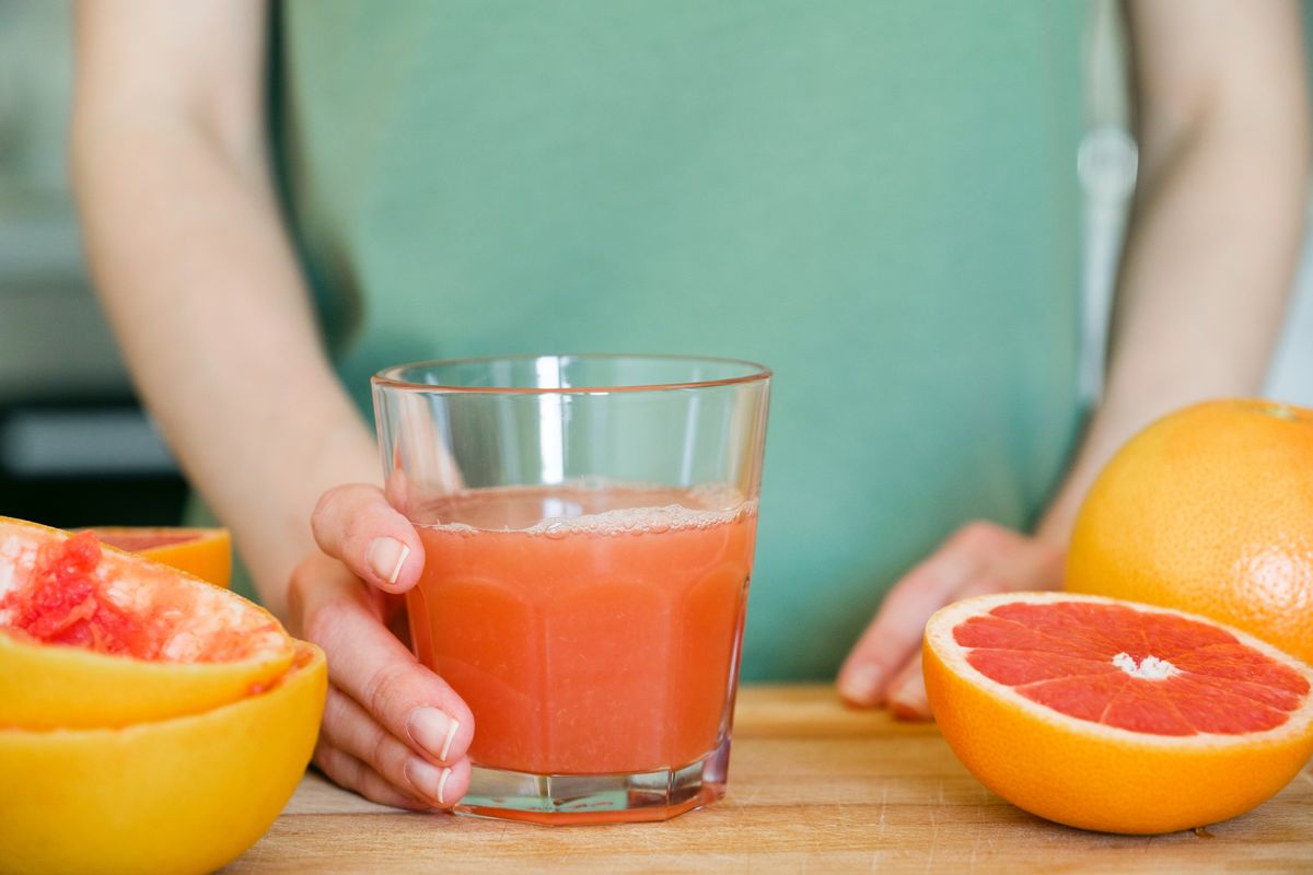 A girl or a woman is holding a glass of freshly squeezed juice from a red ripe grapefruit, on a cutting board, against the background of a wooden kitchen table. I make juice for breakfast. The concept of vegetarian, vegan and raw food.