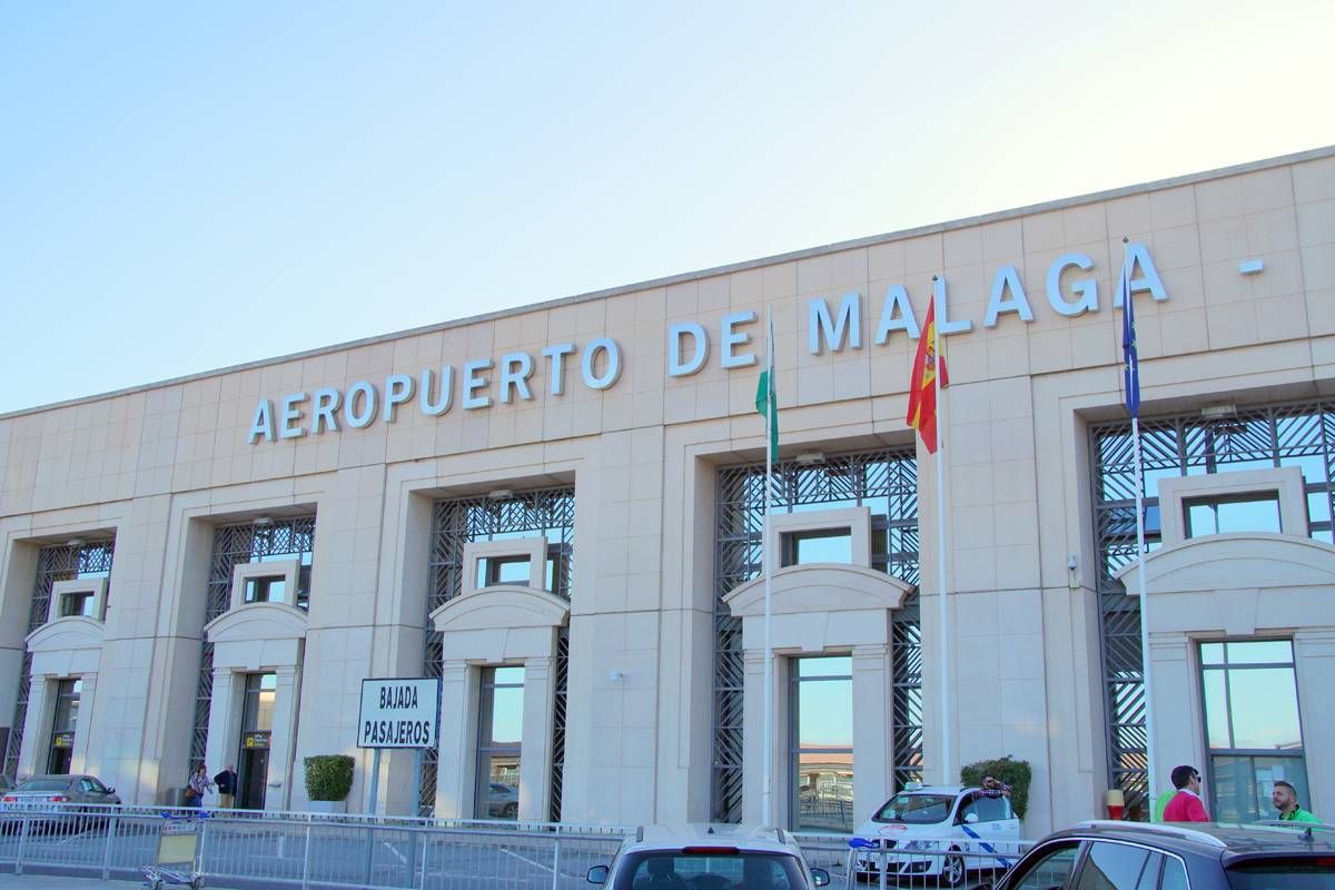 The image features the exterior of a modern building, prominently displaying the words "Universidad de Málaga" above its entrance. The building has a contemporary architectural design with large glass windows and pillars supporting the structure. Several flags are mounted on the side of the building, indicating a sense of national or institutional pride. In the foreground, several cars are parked in front of the building, suggesting a busy environment. The sky is clear, and the lighting highlights the building's features.