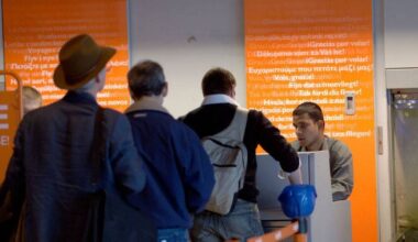 An adult woman with short blonde hair, wearing a navy blue shirt, stands smiling in a modern kitchen setting. The background includes a white wall adorned with a decorative item and a kitchen appliance partially visible.