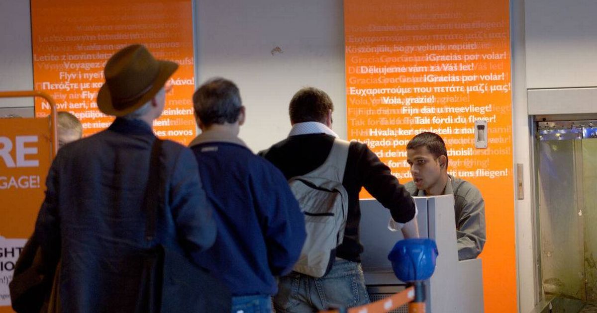 An adult woman with short blonde hair, wearing a navy blue shirt, stands smiling in a modern kitchen setting. The background includes a white wall adorned with a decorative item and a kitchen appliance partially visible.