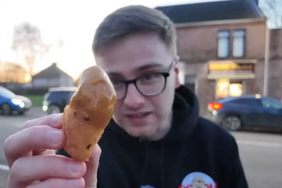A person wearing glasses and a black hoodie is holding up a piece of fried food, possibly a doughnut, in front of a building with parked cars on the side of the road.