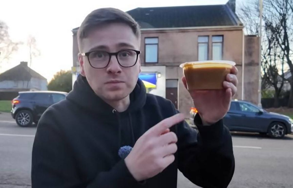 An individual dressed in a black jacket and glasses is standing on the street, gesturing towards a cup of beverage in their hand. Behind them, a building with multiple windows is visible, and in the background, several cars are parked.