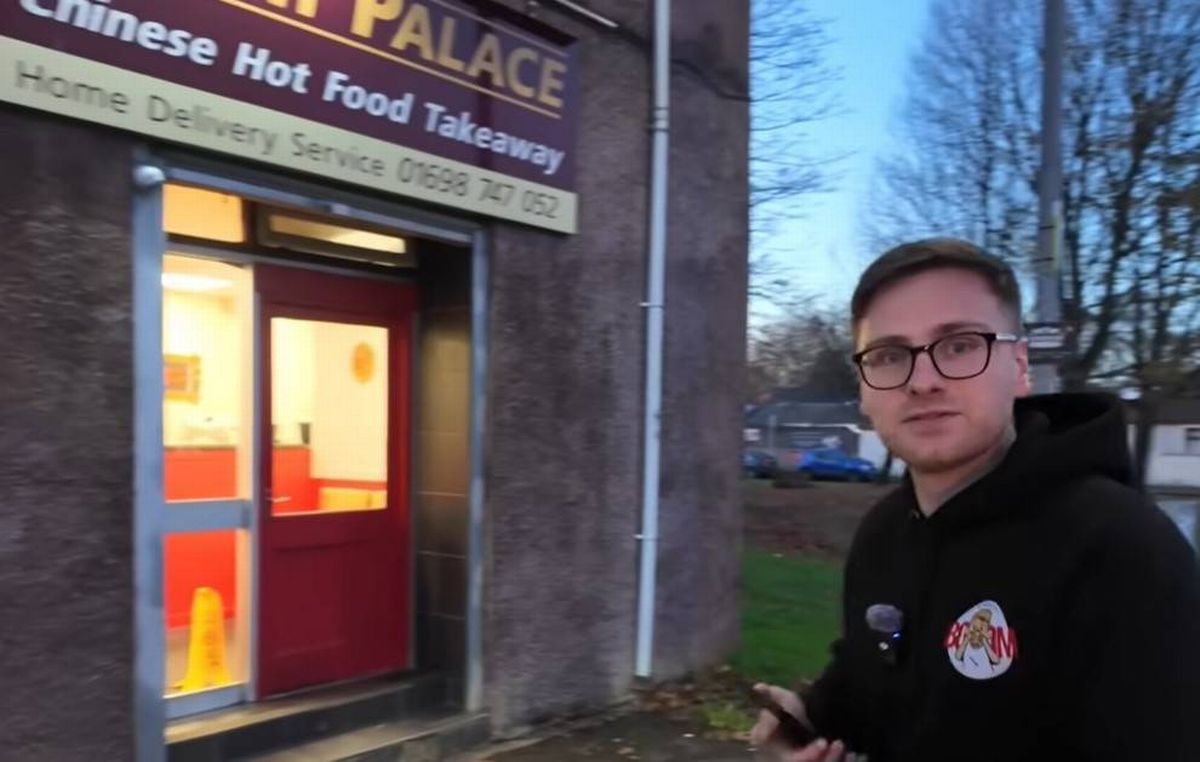 An individual wearing a black jacket and glasses stands outside a commercial establishment, "Chinese Hot Food Palace," with a red door and illuminated signage, set against a backdrop of bare trees and a clear sky.