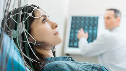 yacobchuk/Getty Woman lying on a medical bed wearing a cap with multiple electrodes and wires attached to the head. In the background, a healthcare professional in a white coat is examining brain scan images displayed on a monitor.