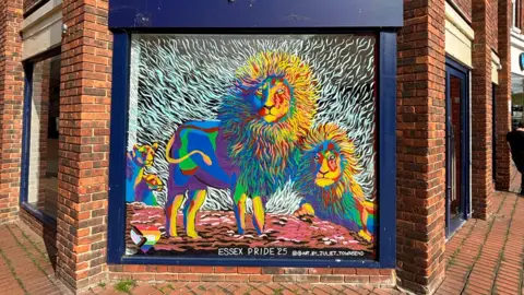 Art by Juliet Townsend Colourful rainbow lions have been painted on a window in a high street. The frame of the window is navy blue and the bricks around it are brown. 