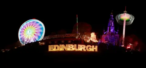 Harris Miller-Saleem The word Edinburgh lit up on a big sign with a ferris wheel and another fairground attraction at either side. The photo was taken at night.