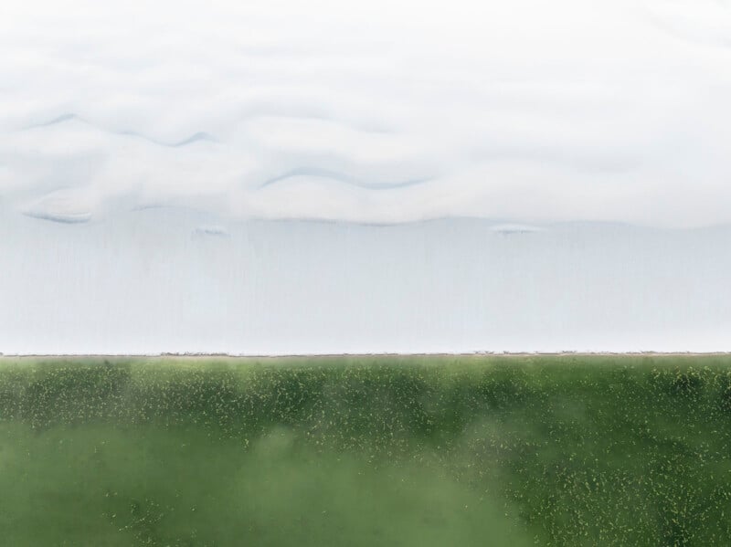A minimalist aerial view of a snowy landscape meeting a green field, with a soft, cloudy sky blending into the white horizon above the stark boundary line.