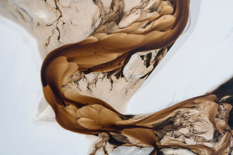 Abstract aerial view of a winding brown river or sand formation curving through a pale, textured landscape with swirling patterns and contrasting light and dark tones.
