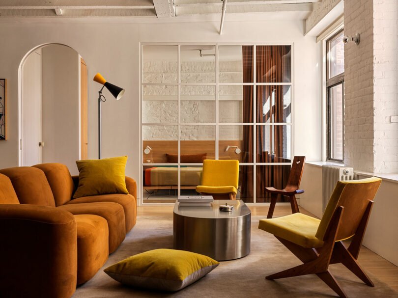 Modern living room with rust and yellow furniture, a round metal coffee table, and large windows; a glass partition separates another seating area with a daybed 