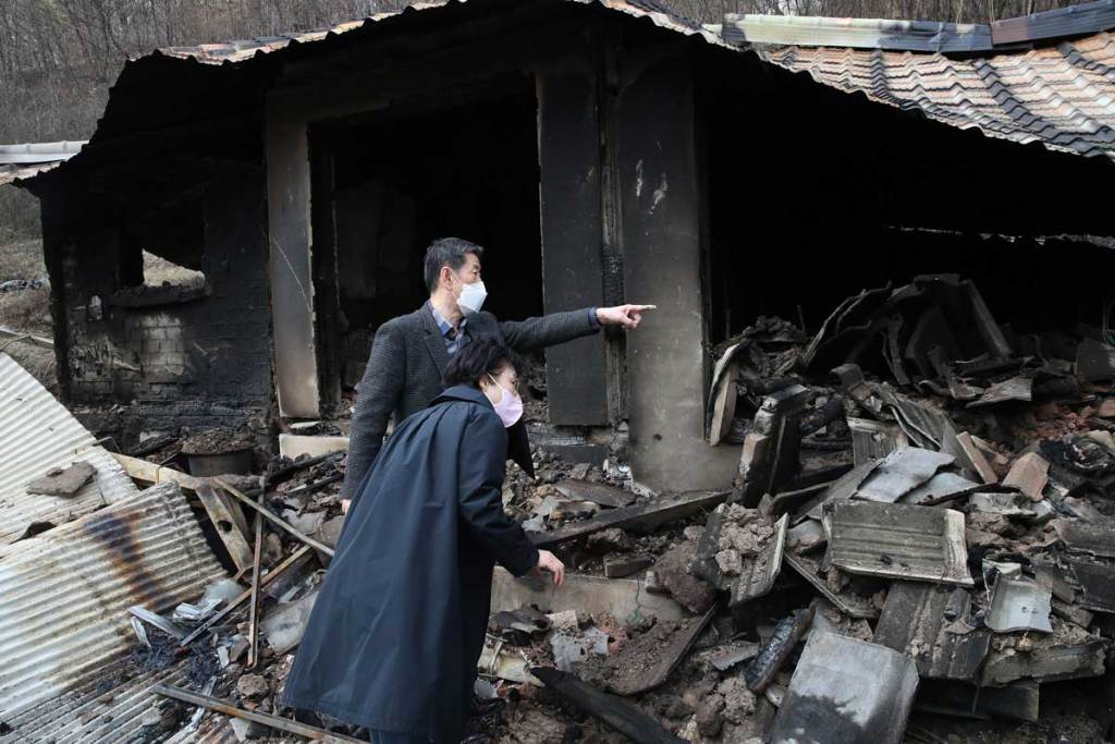 Two people stand in the ruins of a burned home. They're both wearing KN94-style masks.