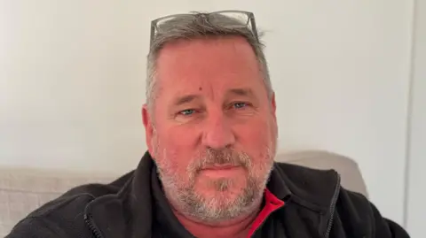 George King/BBC A head and shoulders image of Peter Lenney. He is wearing a black jacket over a black polo shirt. He is looking directly into the camera.