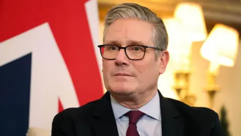 EPA Headshot of Sir Keir Starmer in a suit sitting in front of two bright lamps and a UK flag.