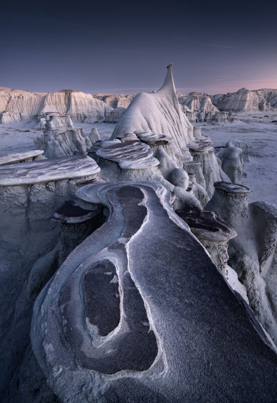 Eroded rock formations and layered stone slabs create a surreal, otherworldly landscape under a dusky sky in a barren desert region.