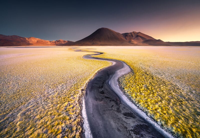 A winding, dark stream cuts through a vast, yellow and white salt flat, leading toward distant mountains under a dramatic sunset sky with deep blue and orange hues.
