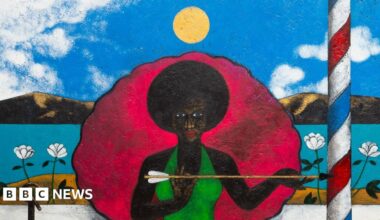 Madame La Sirene by Abe Odedina it depicts a black woman in a green dress holding an arrow and sitting in an large clam shell with a striped barbers pole beside her. In the foreground is a white and black dog in an orange and black basket on a red rug.