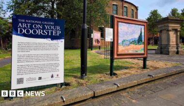 An interpretation board next to a replica artwork. The board has a dark blue top with white writing that reads The National Gallery Art on Your Doorstep. The artwork and board have been installed on a patch of lawn