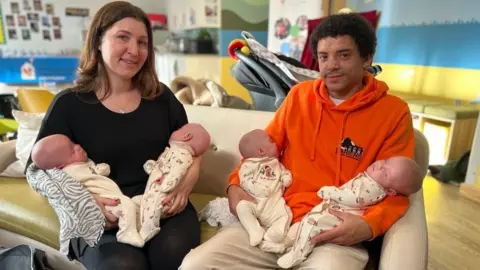 BBC Jodie and Lyde pose with their babies - two each - on a sofa in Ronald McDonald House. Jodie wears a black top and trousers, Lyde an orange hoodie and beige trousers.