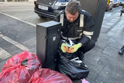 An environmental officer wearing dark protective clothing and clothes reads a scrap of paper taken out of a black bin liner
