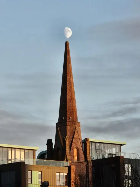 Neil Moir A red-brick church spire with the moon seen directly above it. 