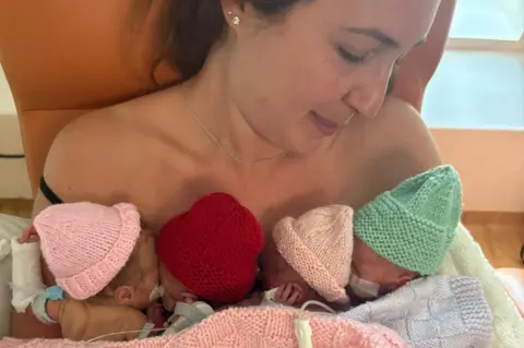 Jodie Keeley Jodie holds the four little babies to her chest during their time in care. They have assorted wires connected to them. They wear knitted hats and knitted blankets are around them.