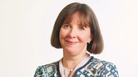 Public Health Agency A professional photograph of Dr Joanne McClean. She has a short hairstyle, cut into a bob. She is smiling at the camera and is wearing a patterned cardigan and a pale pink shirt. She is standing in front of a light background.