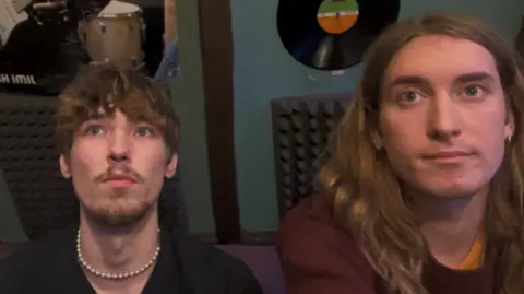 Two young men, one with longer hair sat on the right, sat together in a music studio with records, sound insulation and posters on the wall behind them