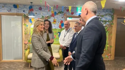 The Duchess meets with officials inside the facility. She is wearing a grey suit. There are colourful paintings of balloons and animals on the wall, one looks like Pooh bear.