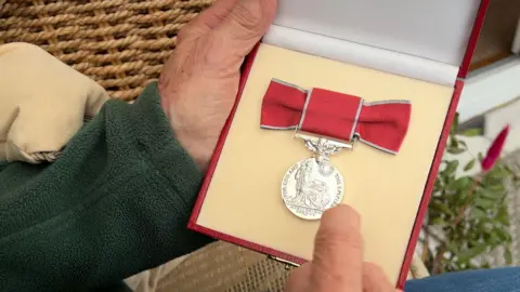 Joan Lockley sits in a brown wicker chair and holds her British Empire Medal in her hands. The medal is a silver circle engraved with a figure of Britannia wearing robes and carrying a sword and spear. Around the image are the words "For God and the empire, meritorious service"