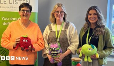 Sarah Kelly from Manx Lottery Trust and Isle Listen's Becks Macnair and Holly Ramsay stand behind the table smiling for the camera. They are holding stuffing children's toys used in the lessons.