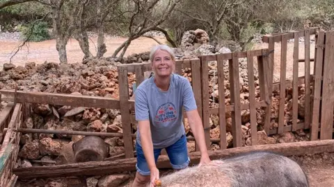 Menorca Donkey Rescue Jane White is wearing a grey t shirt which says 'it's time to listen,' on it in blue writing. She is wearing blue denim shorts and is stood up with her hands on the back of one of the pigs at the sanctuary. 