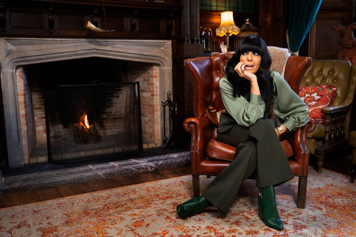 Claudia Winkleman sat down on a chair next to a fireplace, resting her arm on her knee.