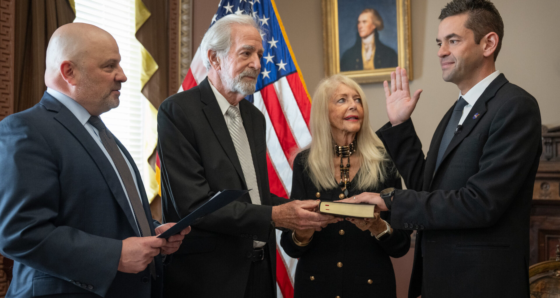 Four people, including NASA Administrator Jared Isaacman, stand in an office with an American flag in the background, as Isaacman is sworn in, with his right hand raised.