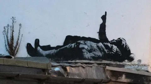 Banksy The black and white street art depicts two children lying on their backs and pointing upwards. The child nearest the camera is wearing a wooly hat with hands tucked into their jacket pockets. Behind them a larger person is pointing upwards in a bobbly hat (possibly a Santa type hat) also wearing boots.