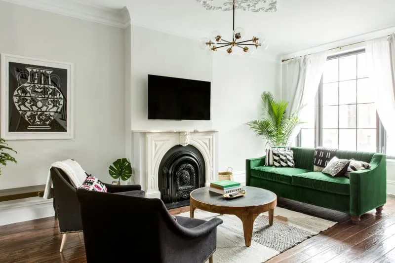 Living room with green velvet sofa, black armchairs, white fireplace, round coffee table, and large window with sheer curtains.