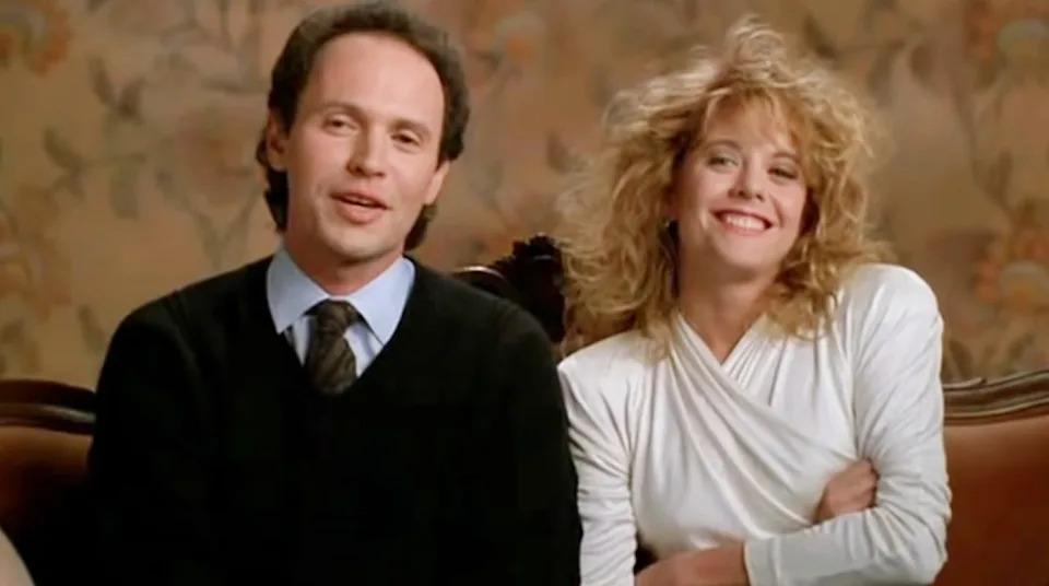 A man and woman sit on a sofa. The man wears a suit and tie, and the woman wears a white top with tousled hair, both smiling warmly