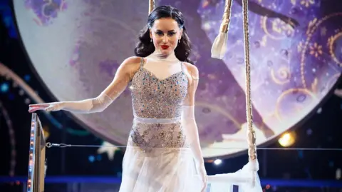 PA Media Amber Davies pictured in one of her Strictly performances. She is wearing a silver, sequinned dress and is too on a platform. The Set design in the background shows a full moon.