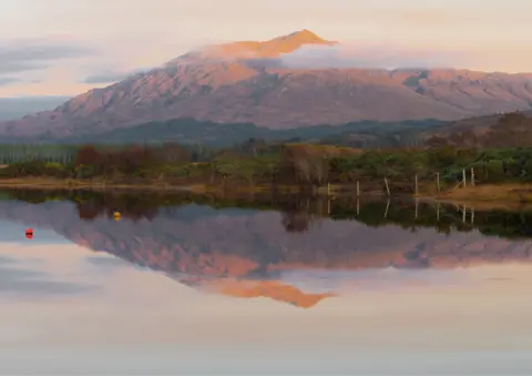 Jacqueline Shaw A mountain with wisps of cloud near the top. The mountain is reflected in the water.

