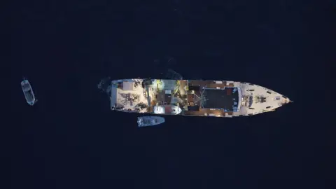 James Glancy/Martin Stalker The image is captured by a drone and is looking directly down upon a research vessel - with crew and equipment visible on deck - and two smaller dinghies close by. The picture is taken at dusk and the sea surrounding the vessel looks dark, inky blue